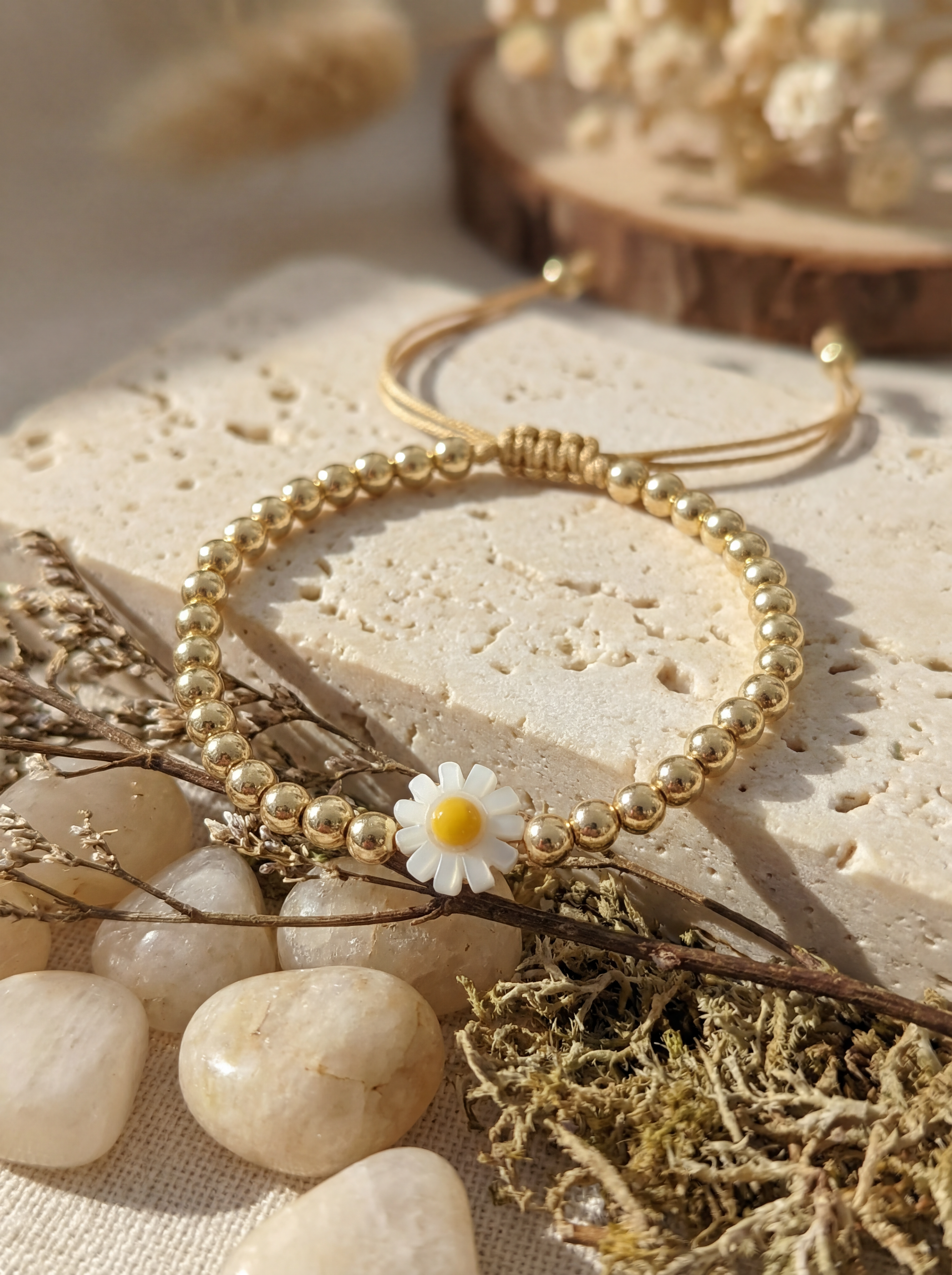 Gold beaded bracelet with a daisy charm on a natural stone surface with dried plants.