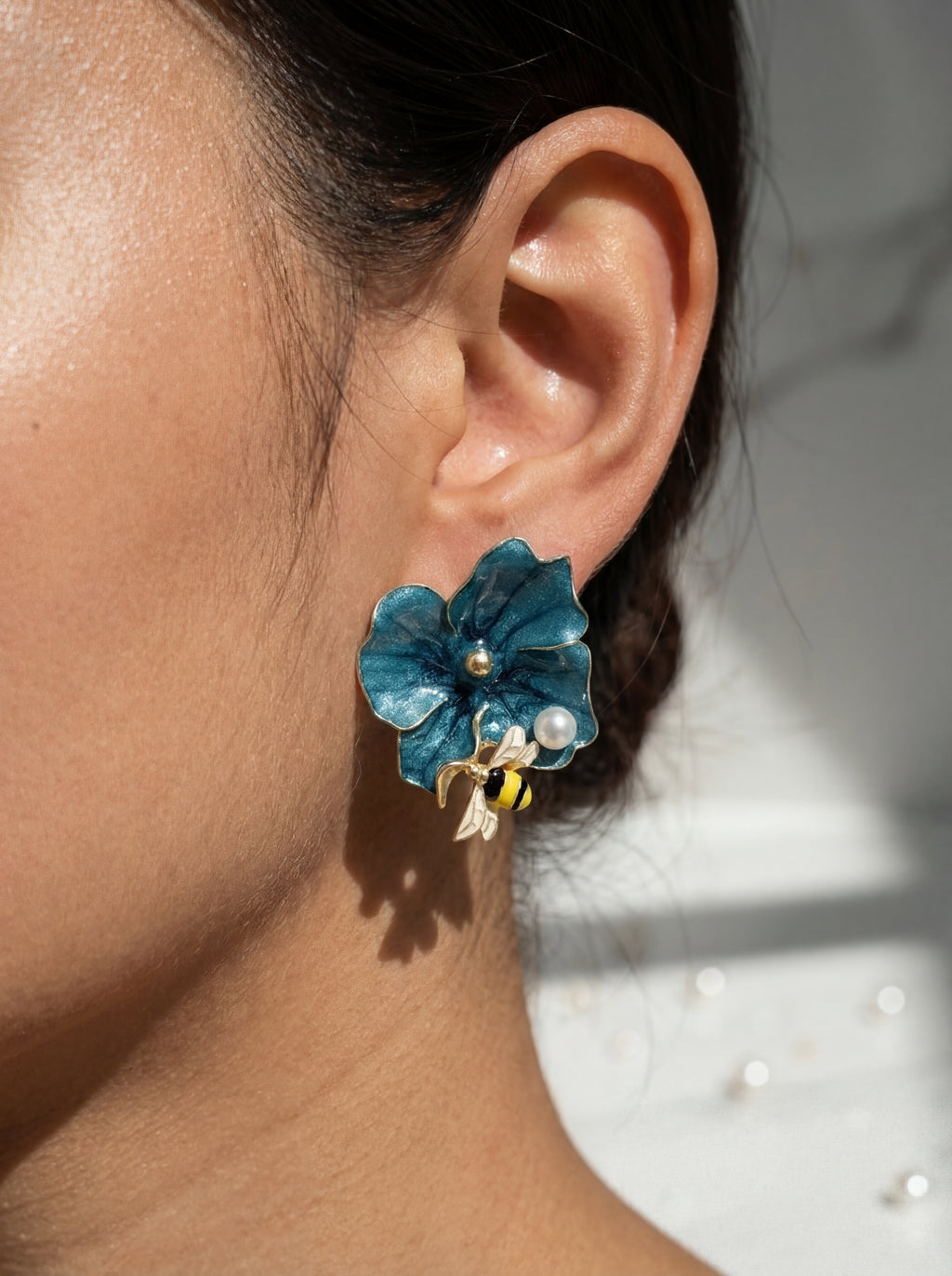 Close-up of an ear wearing a blue floral earring with a bee and pearl detail.
