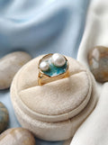 Gold ring with pearls and blue stone on a beige cushion, surrounded by stones.