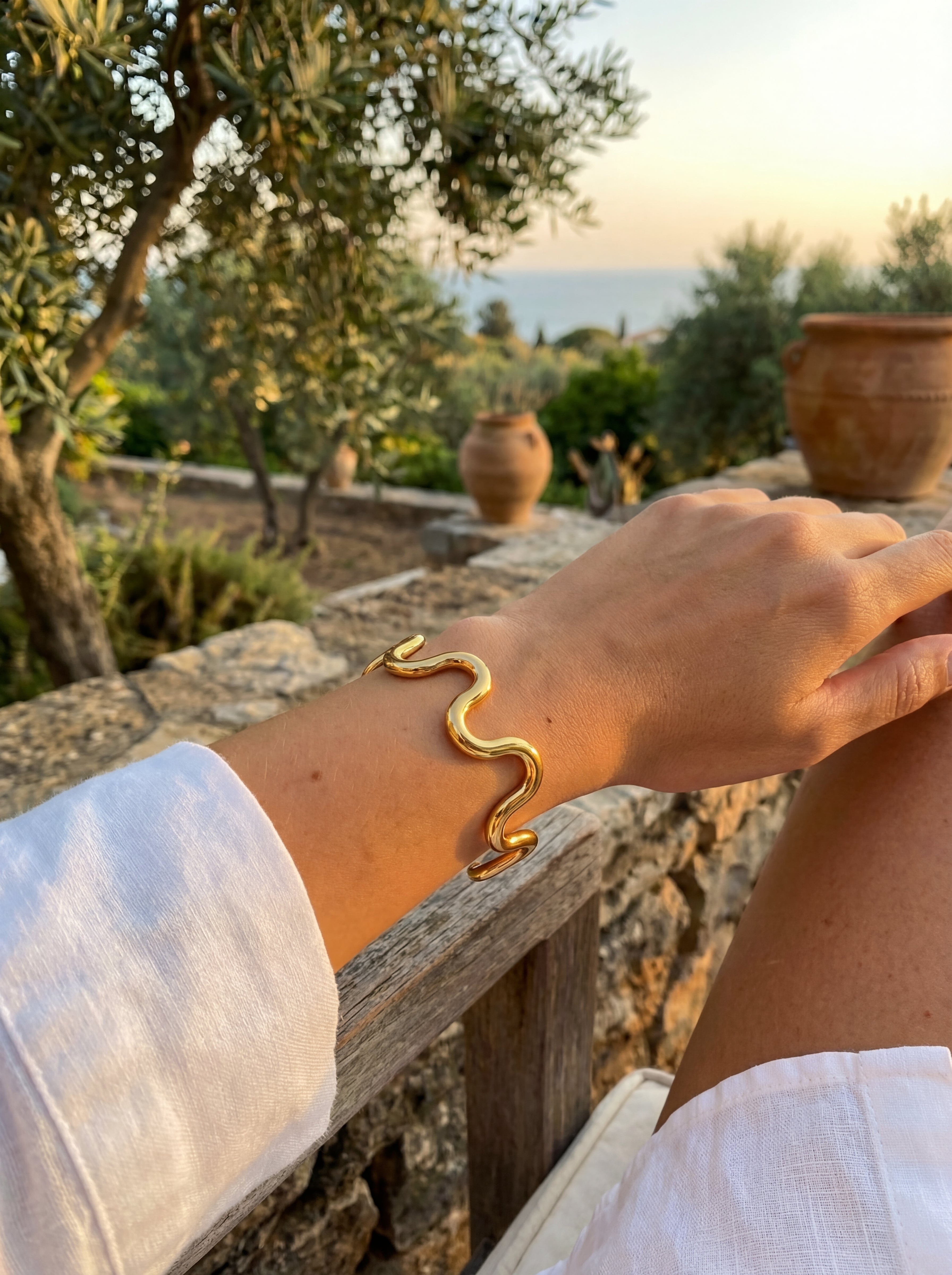 Gold bracelet on a wrist with a scenic background of trees and pots.