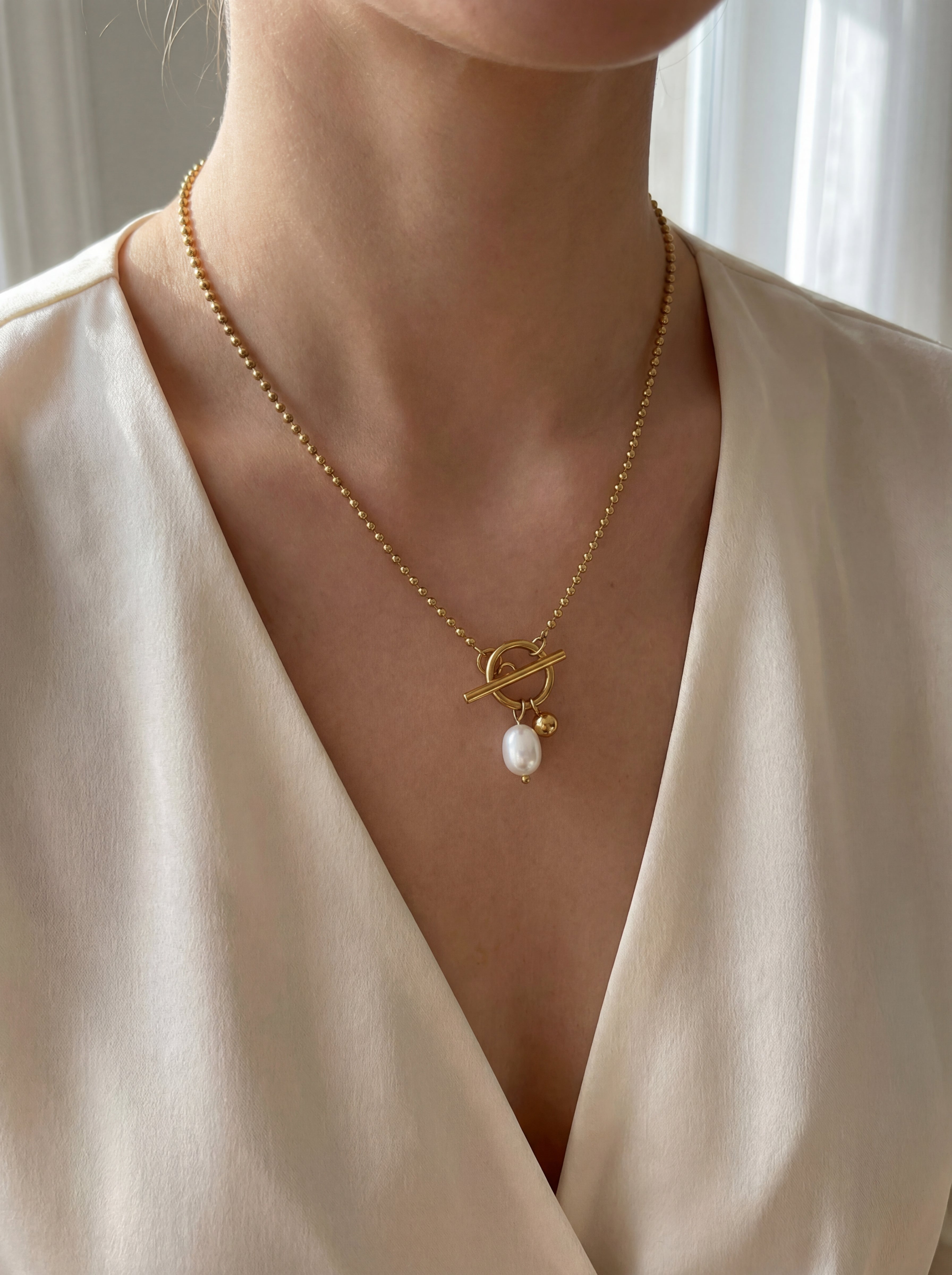 Gold necklace with a pearl pendant worn by a person in a white outfit.