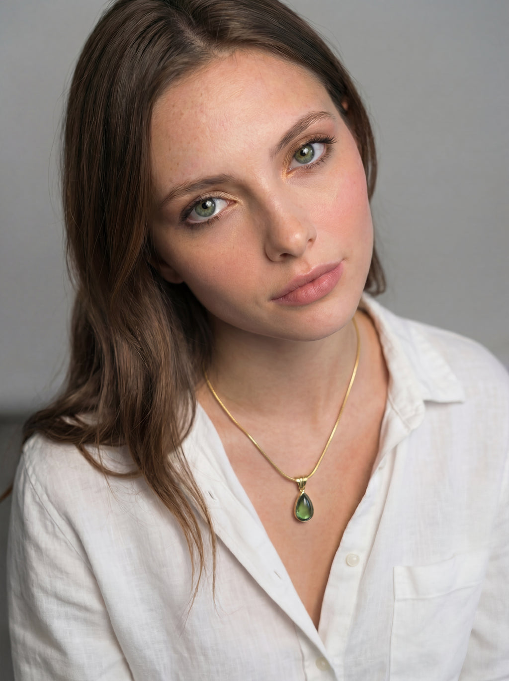 Woman wearing a white shirt and gold necklace with a green pendant against a gray background