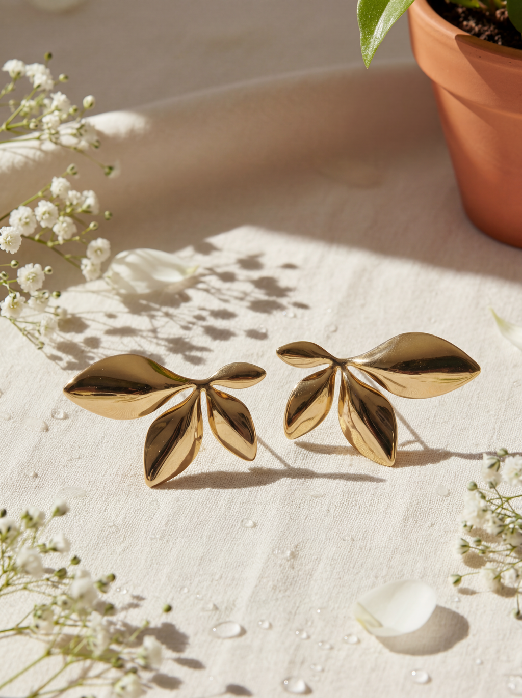 Gold leaf-shaped earrings on a light surface with white flowers and a plant pot in the background.