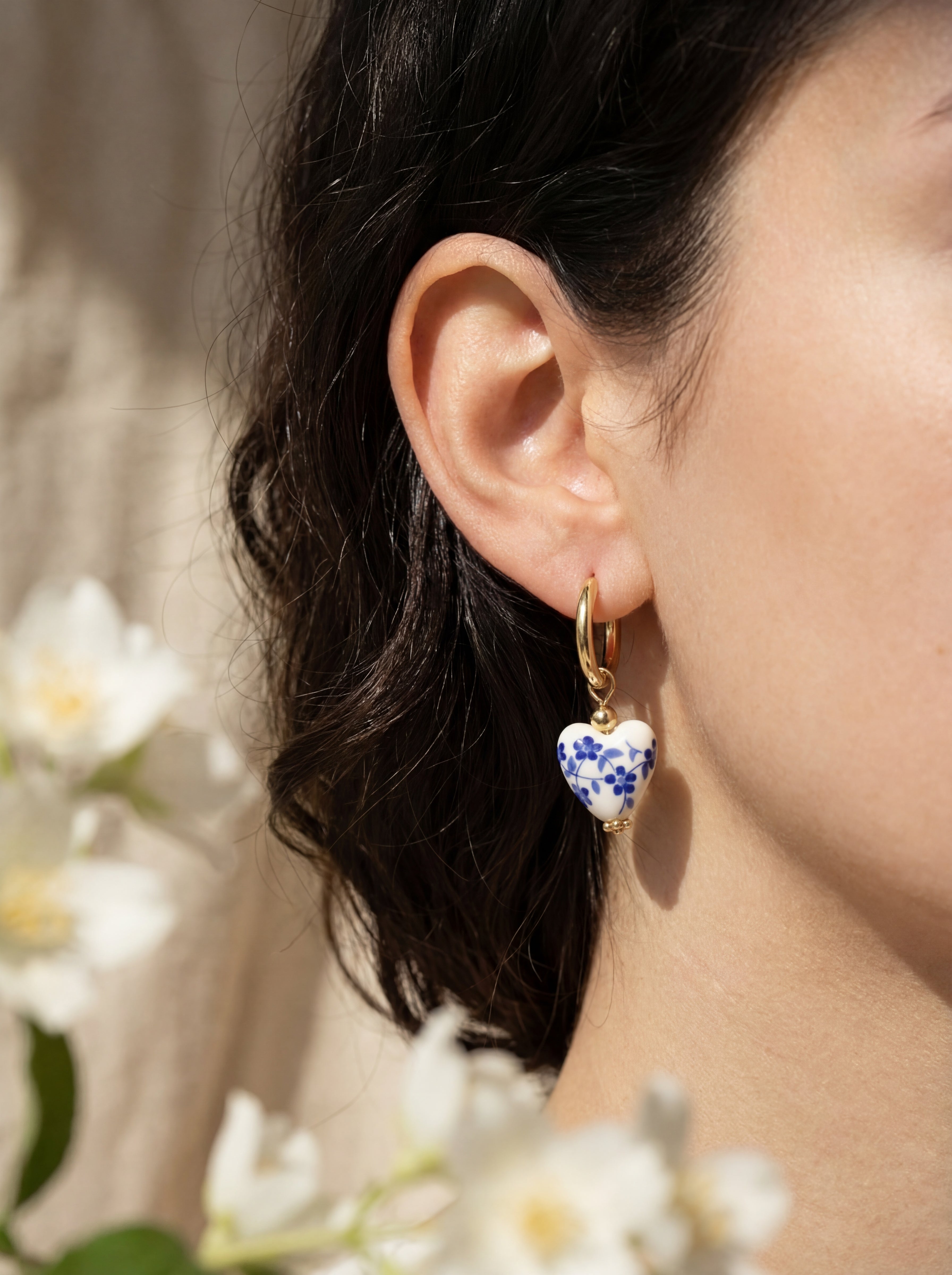 Close-up of an ear wearing a heart-shaped earring with floral design, surrounded by white flowers.