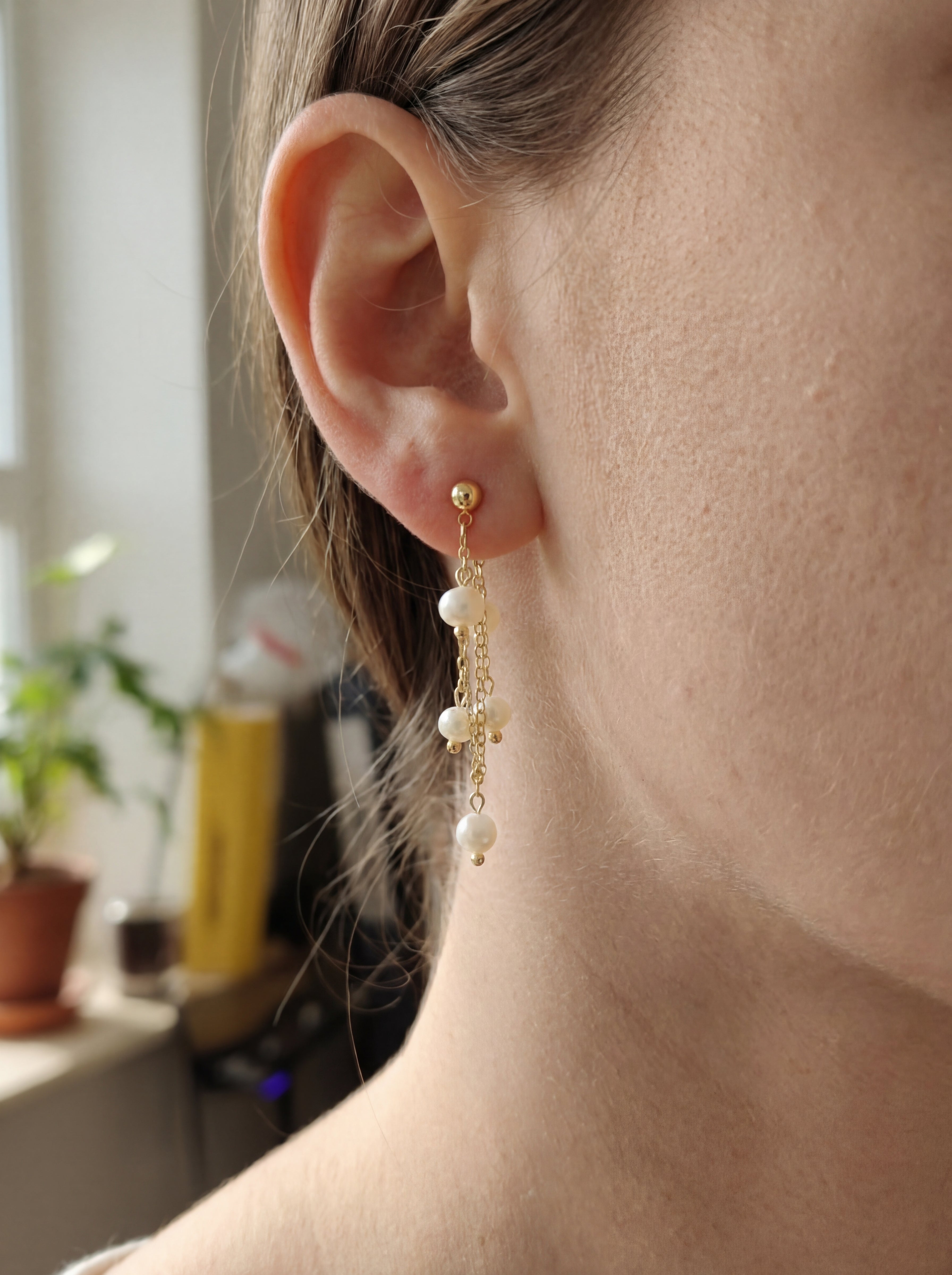 Close-up of a person wearing gold earrings with pearl drops, blurred indoor background.
