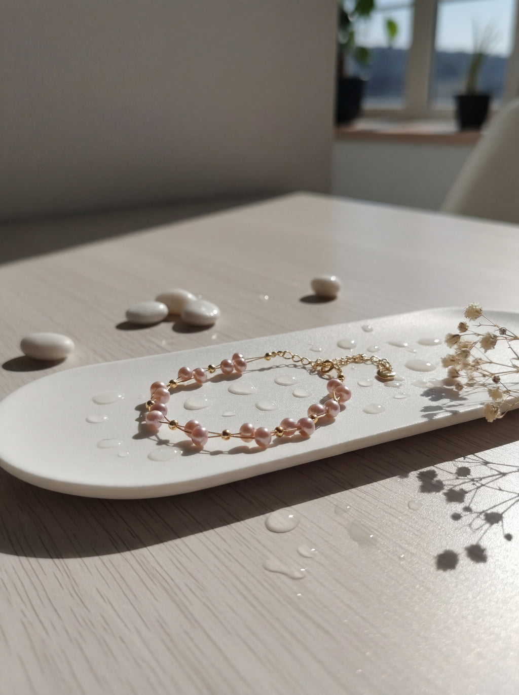 Pearl bracelet on a white plate with small stones and dried flowers on a light wooden surface.