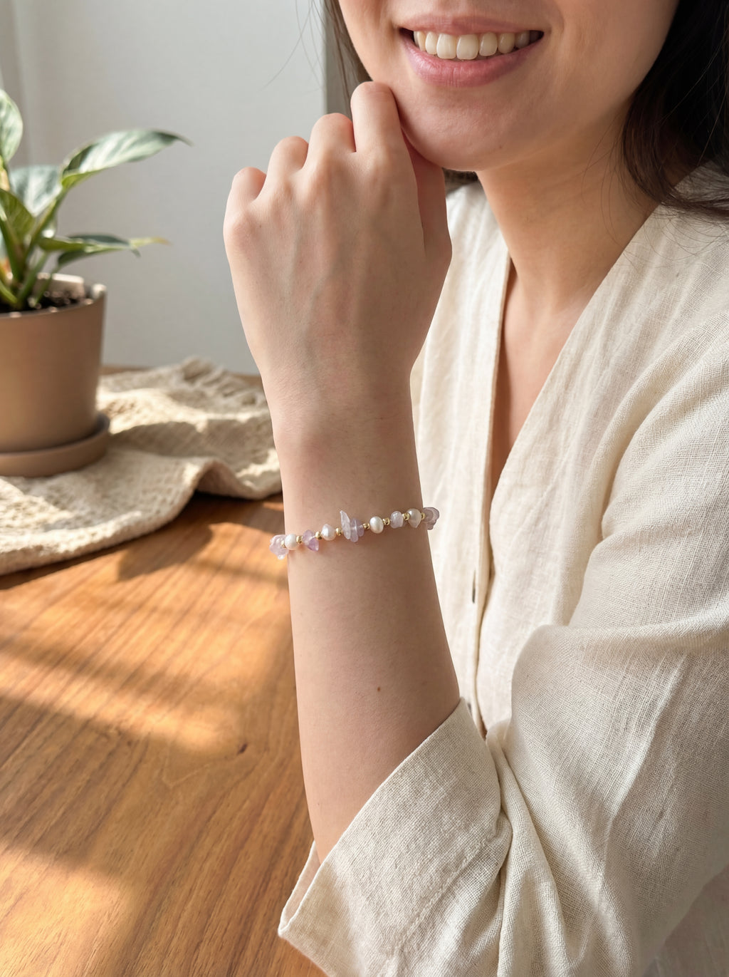 Woman wearing a pearl bracelet in a home setting with a plant and wooden floor.